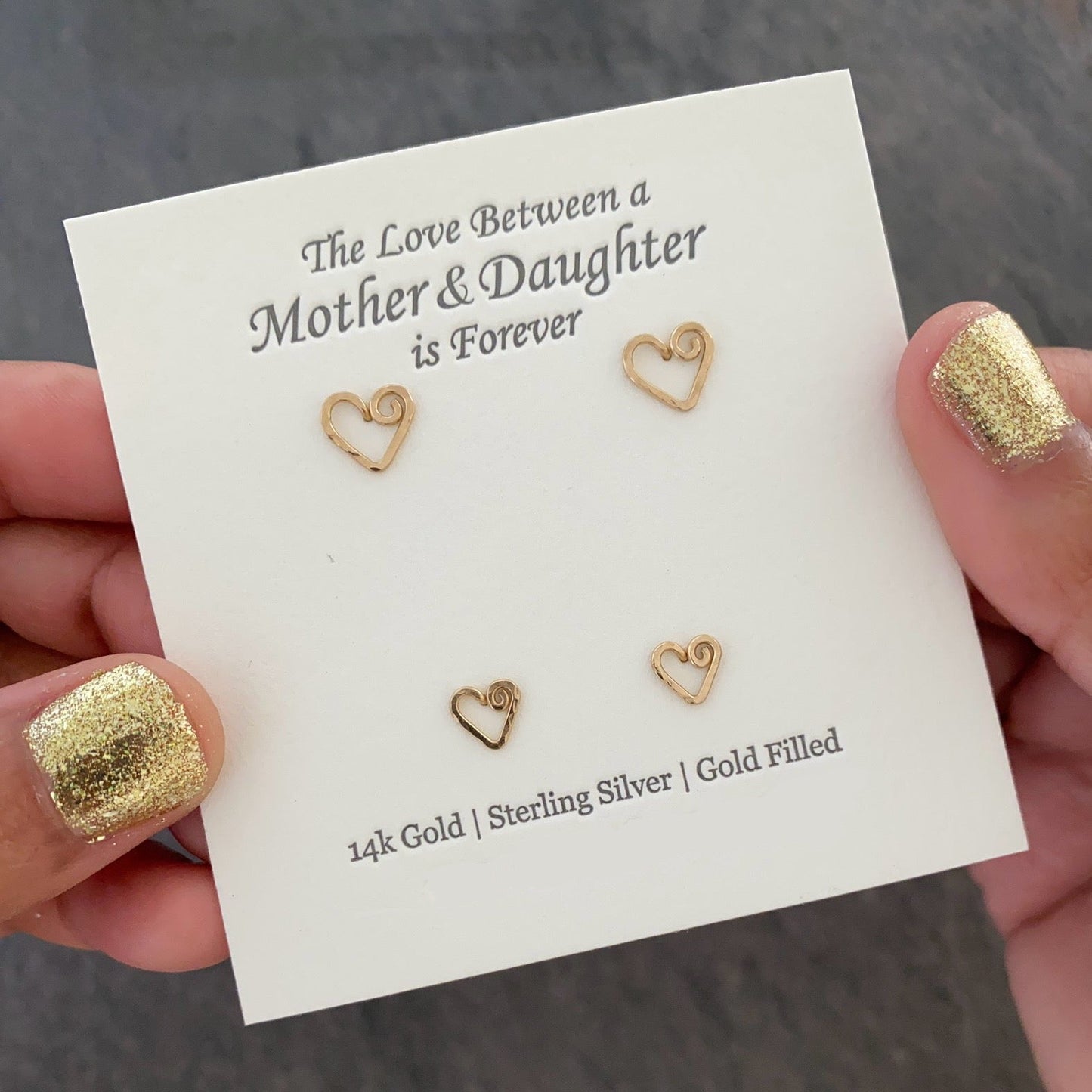 Gold heart-shaped earrings on a card with text held by hands with gold nail polish.