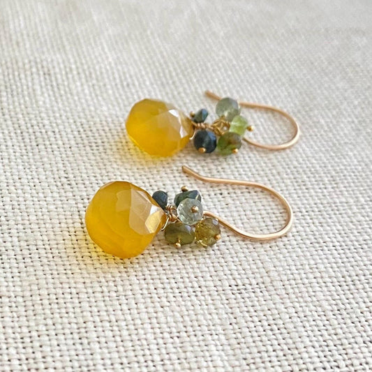 Pair of gold earrings with yellow beads and green gemstones on a textured fabric background